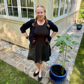 Woman wearing Birdie & Claire Stephanie Dress in Forest Green and Dana Cropped Sweater in Navy standing outdoors near a house with potted plants.