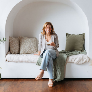 Woman sitting on a white bench holding a cup of coffee with green cushions in a cozy room. Wearing Birdie & Claire Elise Sweater in cement color.
