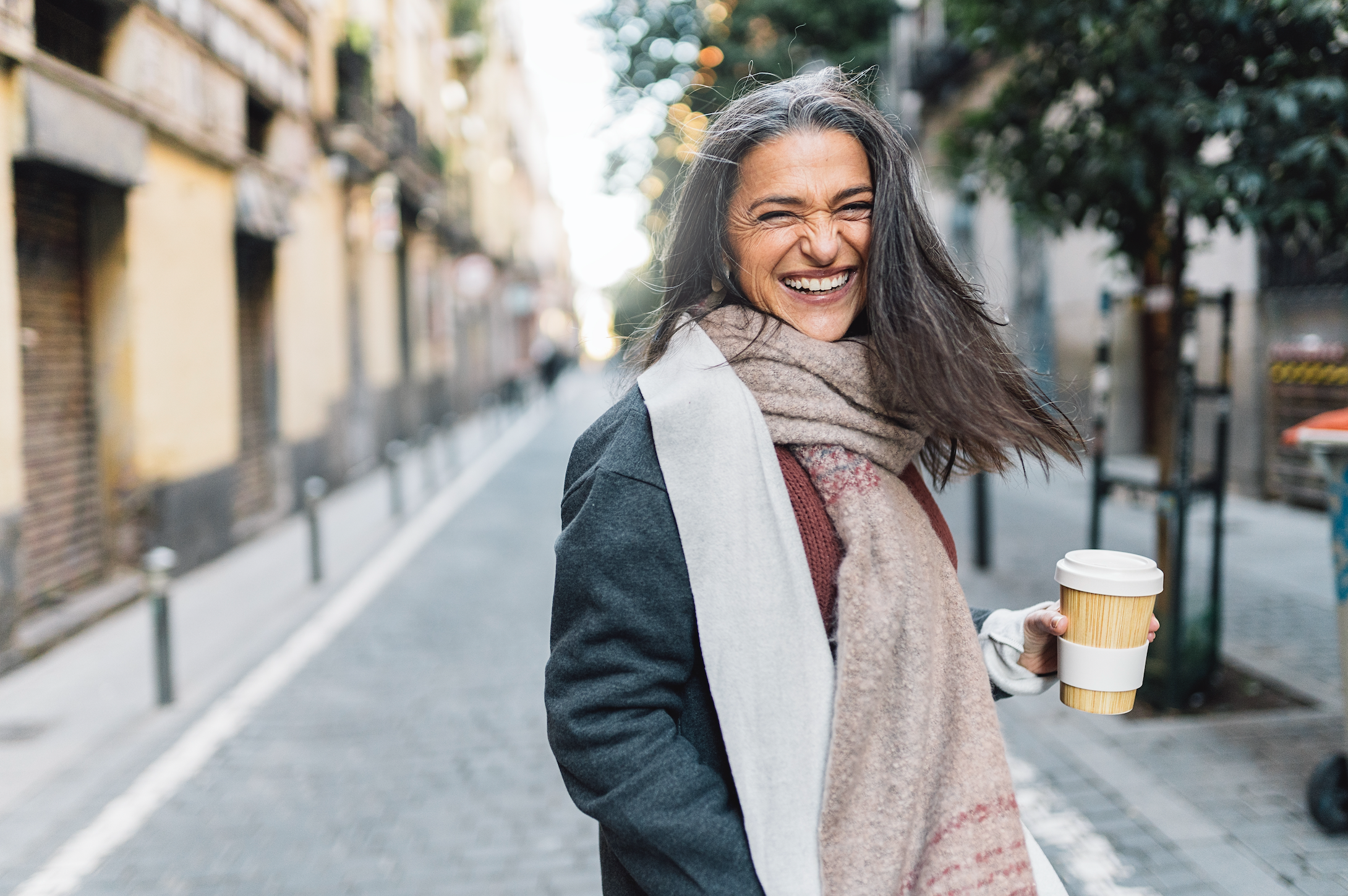 Woman in jacket and scarf, smiling outside and holding coffee cup